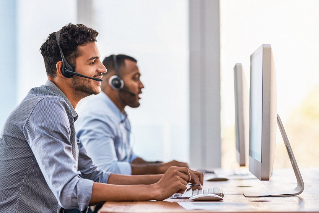 Two men sitting at desktop computers talking on the phone